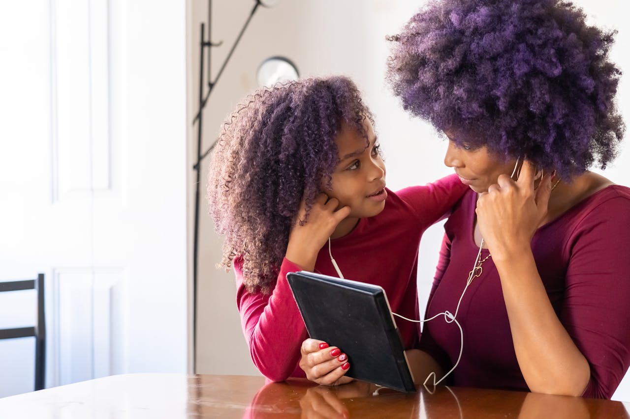 Mujer adulta y niña compartiendo audífonos conectados a una tableta mientras se miran. Ambas visten similar y son afro.
